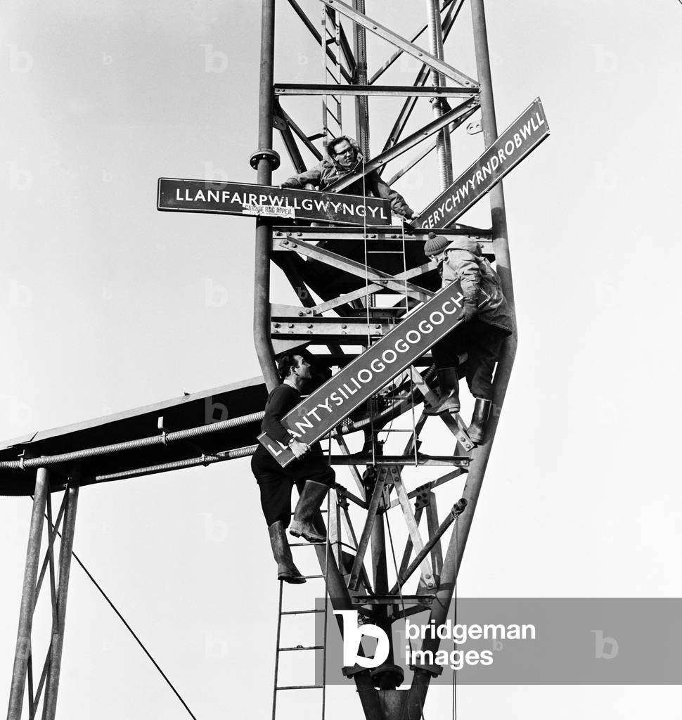 North Wales students removed the very long sign of the Llanfairpwllgwyngyllgogerychwyrndrobwllllantysiliogogogoch railway station in Anglesey and carried it to South Wales before hositing up onto the Wenvoe television mast at St Lythans, near Cardiff. Pictured are TV engineers removing the sign from the mast. 4th February 1964 (b/w photo)