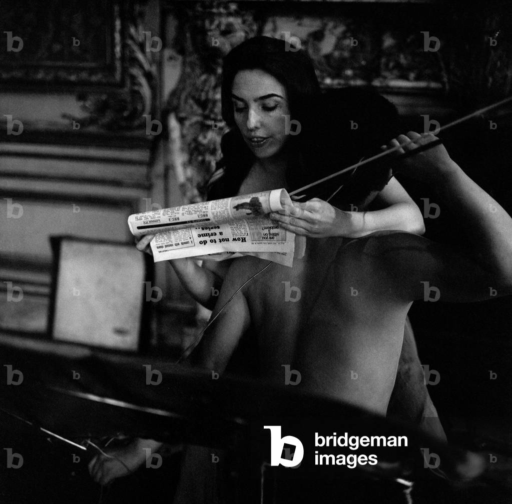 Cellist Charlotte Moorman rehearsing at 18 Carlton House Terrace for her particular sound effect concert. With her is her Korean assistant Mr Nan June Paik. Among her sound effects are gongs, chains, bottles, empty beer cans, a piece of plate glass, dustbin lid, a hot-plate, electric light bulbs, a full coca cola bottle, axe, door and indian bells, butter for sizzling, ancient cymbals, a siren, toy guns, masks, human cello, amplifier, taped and recorded sound, mating call of a duck and her cello. A thing she is short of but is trying to get is the casing of a 100 lb bomb. Pictured is Charlotte reading excerpts from the Daily Mirror across the bare back of her assistant. 22nd September 1968 (b/w photo)