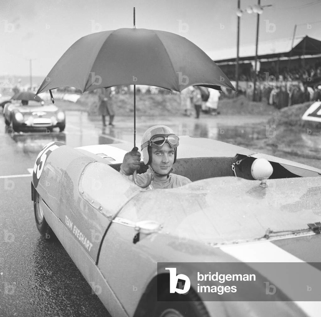 Brian Hart holds an umbrella up to keep off the heavy rain as he waits for the start of the 3 hour race in his Lotus 23b at Snetterton 27th September 1964