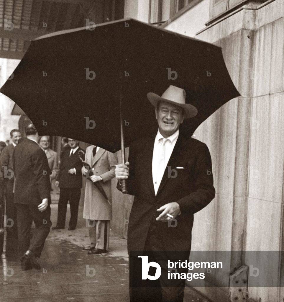John Wayne standing under a big black umbrella in the rain in London, October 1960 (b/w photo)
