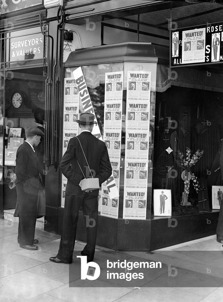 A passer-by look at a tailor shop window plastered with copies of the Daily Mirror Hitler Wanted poster World War II Home Front Shops Gas Mask Box, 4th September 1939 (b/w photo)