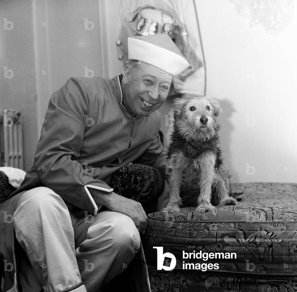 The show must go on, says George Formby, appearing as Mr Wu in 'Aladdin' at the Hippodrome, Bristol. George, who lost his wife at the weekend, relaxes with his pet dog, Punch. 28th December 1960 (b/w photo)