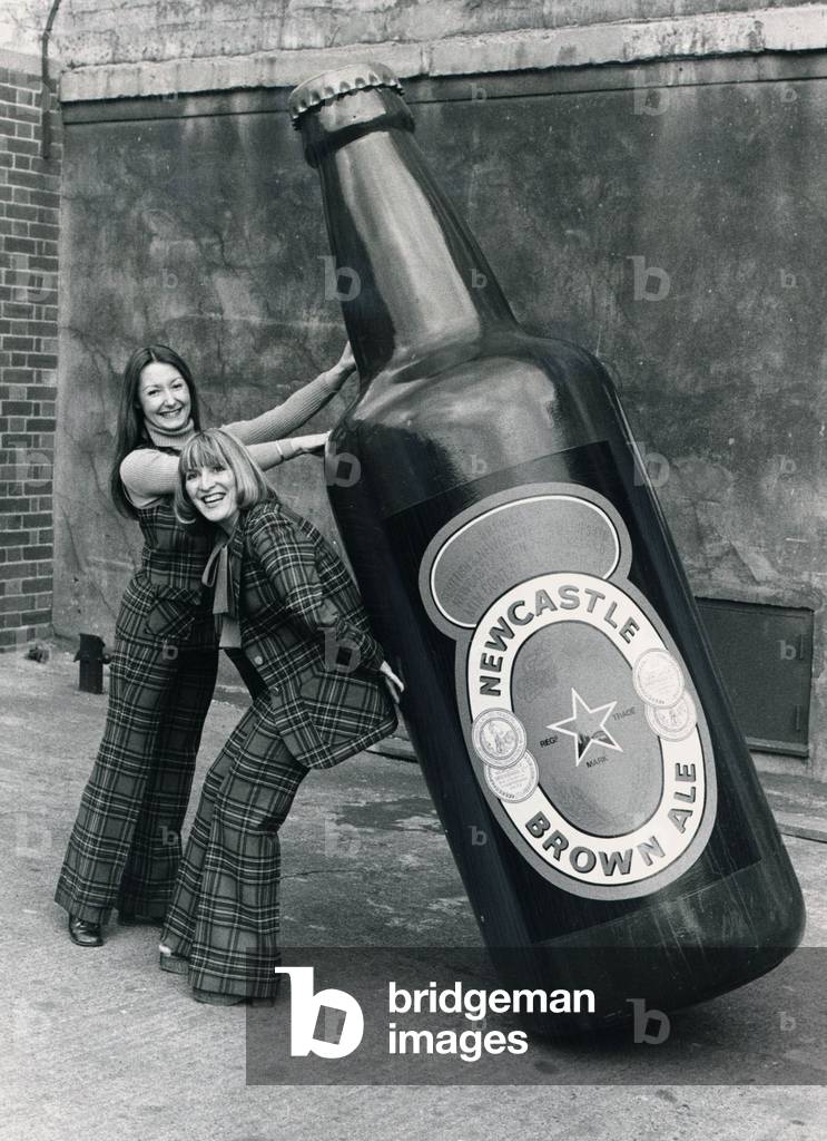 Brenda Kelly (without jacket) and Jennifer Donkin holding a nine foot tall bottle of Newcastle brown ale, c. 1975 (b/w photo)