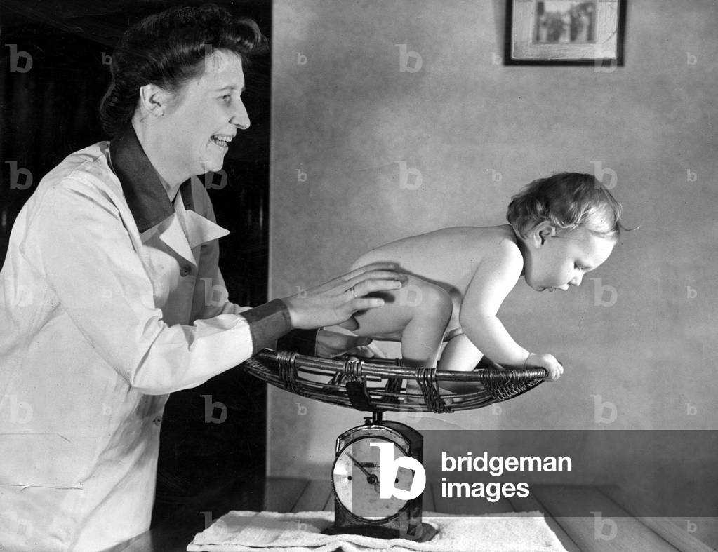 A midwife weighs a toddler in her scales during a check up
July 1943.