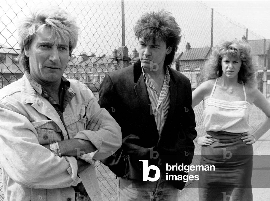 Anyone seen Elton? Puzzled... Rod Stewart and Paul Young with their bodyguard, Libby Krall, in Wimbledon. The singers had got together for a rehearsal for a big gala rock show in aid of the Prince's Trust. But when Elton arrived at the Wimbledon rehearsal rooms, he refused to pose for a picture, 20th June 1986 (b/w photo)