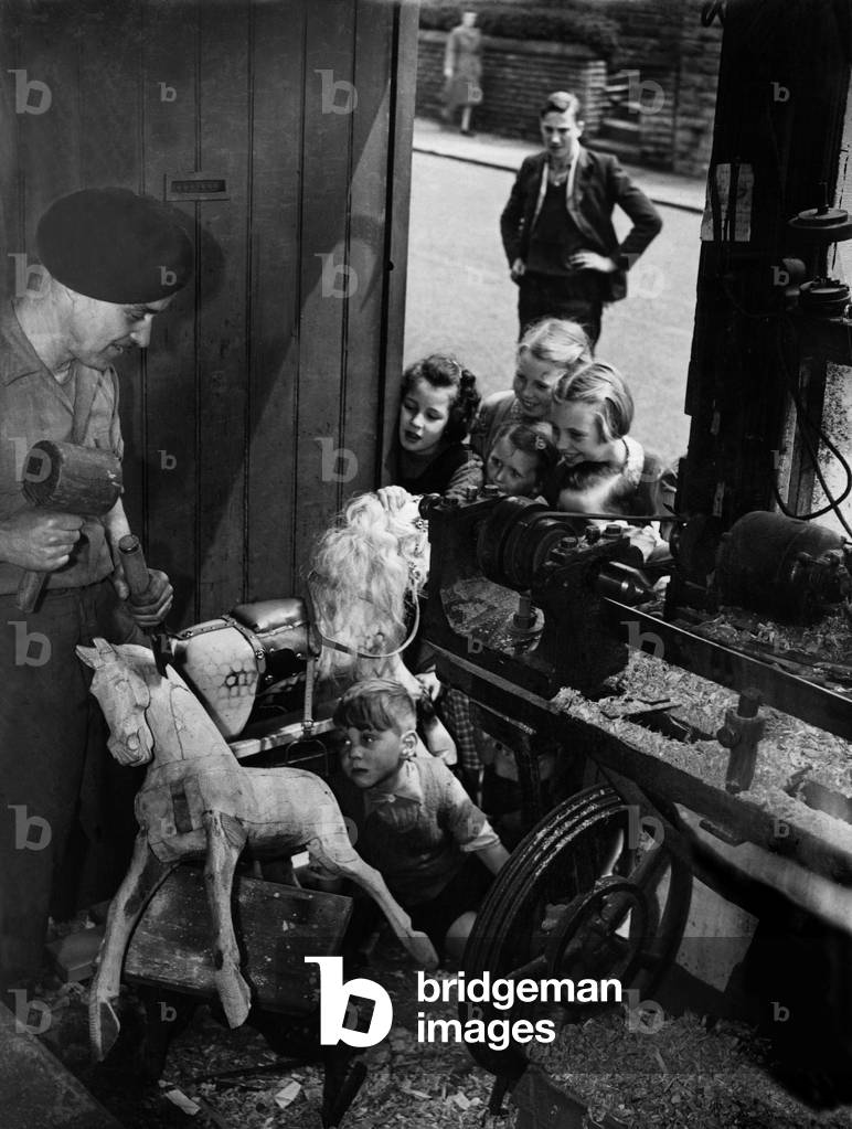 One of the last rocking horse craftsmen in Britain, Steve Winder from Ossett, Yorkshire. Using an axe and chisels he is able to start shaping a horses head in a few minutes. PS Steve in his workshop with an audience of children watching him at work, 22nd May 1949 (b/w photo)