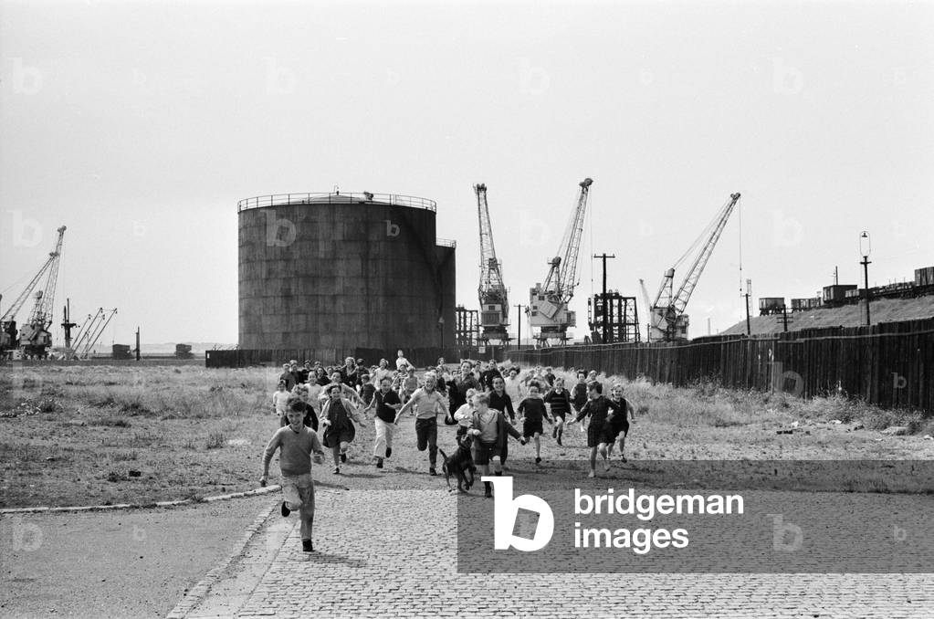 A group of children from Garston, Liverpool enjoy a day out at Sefton Park in the summer holidays. Here, the group rush for the bus to take them to park away from the cranes at Garston docks, pictured in the background. 20th July 1966 (b/w photo)