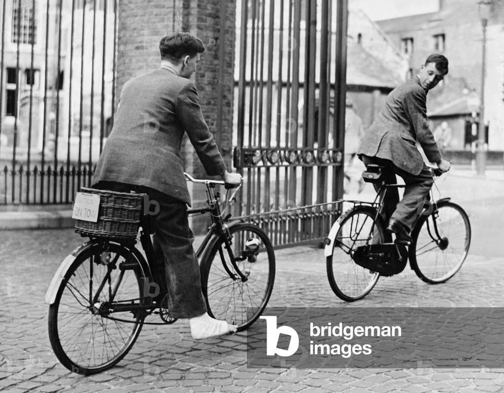 A Cambridge Student with his foot in bandages and sitting on his bike is towed by another pushbike. Sign on bike says Caution - On Tow, May 1949 (b/w photo)