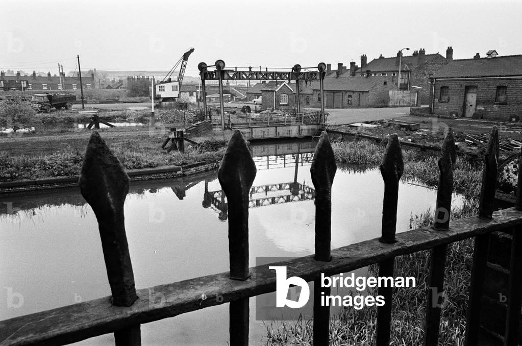 Railings, blackened by 150 years of industrial smoke separate the weed grown banks of the Birmingham Wolverhampton Canal at Factory Locks, Tipton, from a railway coalyard. In the centre is an unusual mid 19th century lifting bridge over the U bend of the canal. Feature on the Black Country, an area of the West Midlands in England, north and west of Birmingham. 25th May 1968 (b/w photo)