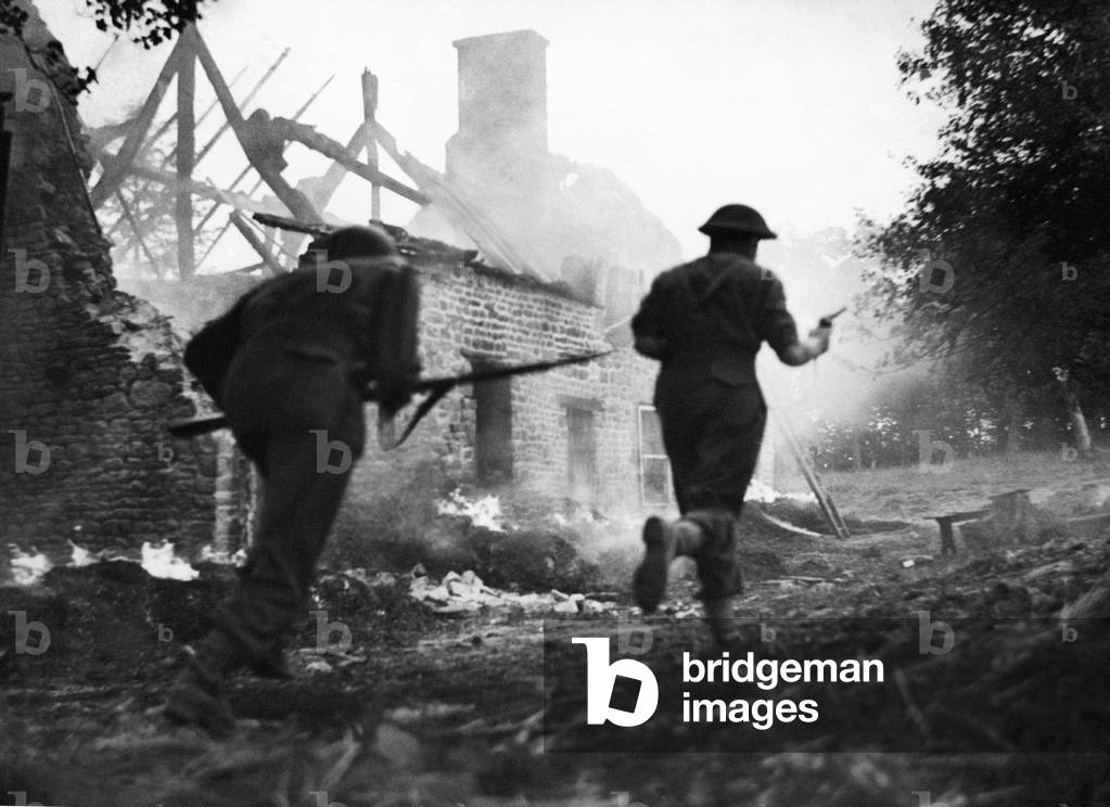 The advance south of Le Beny Bocage continues. British infantry are seen here rushing past the burning remains of a farmhouse in pursuit of the enemy. The Germans had been using the buildings as their quarters. August 1944 (b/w photo)