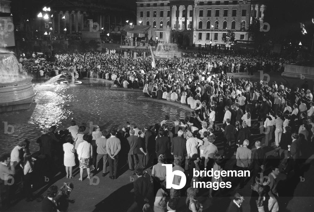 People gather late at night in Trafalgar Square to witness the first Moon landing on a giant tv screen erected for the event. people stand around the fountains and pools, Trafalgar Square London, July 1969 (b/w photo)