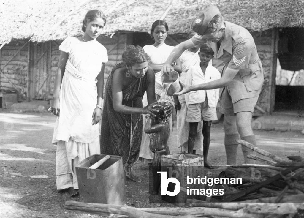 Sergeant Ferris of the Royal Air Force helping to wash the hair of a young child during his round of inspection watched by local Cingalese men and women in Ceylon during the Second World War. 
October 1944