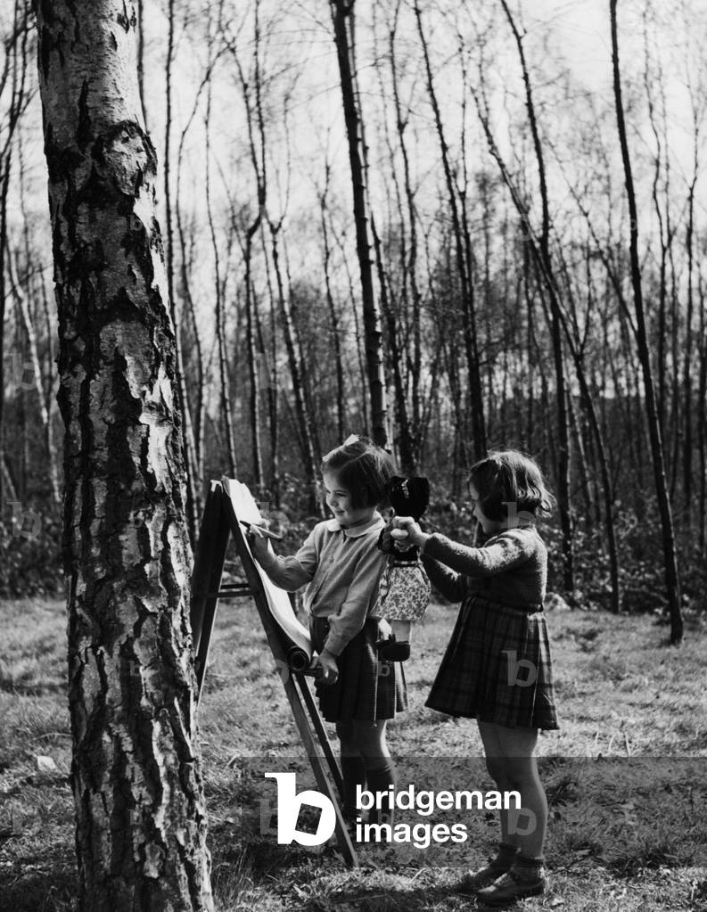 Two girls drawing with crayons and playing with their dolls as they attend their nursery school in Surrey woods. The school is run by the Ministry of Education. 
Circa 1955.