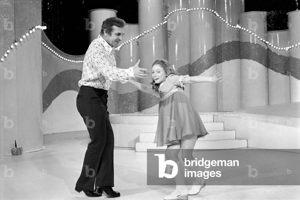 Lena Zavaroni in the Bachelors Show. Lena Zavaroni 10 yrs old has a school lesson in the make up room at the BBC theatre London with the tutor Ellis Gilmer 25 from New Zealand. Lena Zavaroni with Champers a poodle dog in one of the scenes. The Bachelors Lena Zavaroni and director Ernest Maxim. Lena Zavaroni has fun with Bachelor John (with moustashe). A big hug for Lena from Bachelor John. February 1975