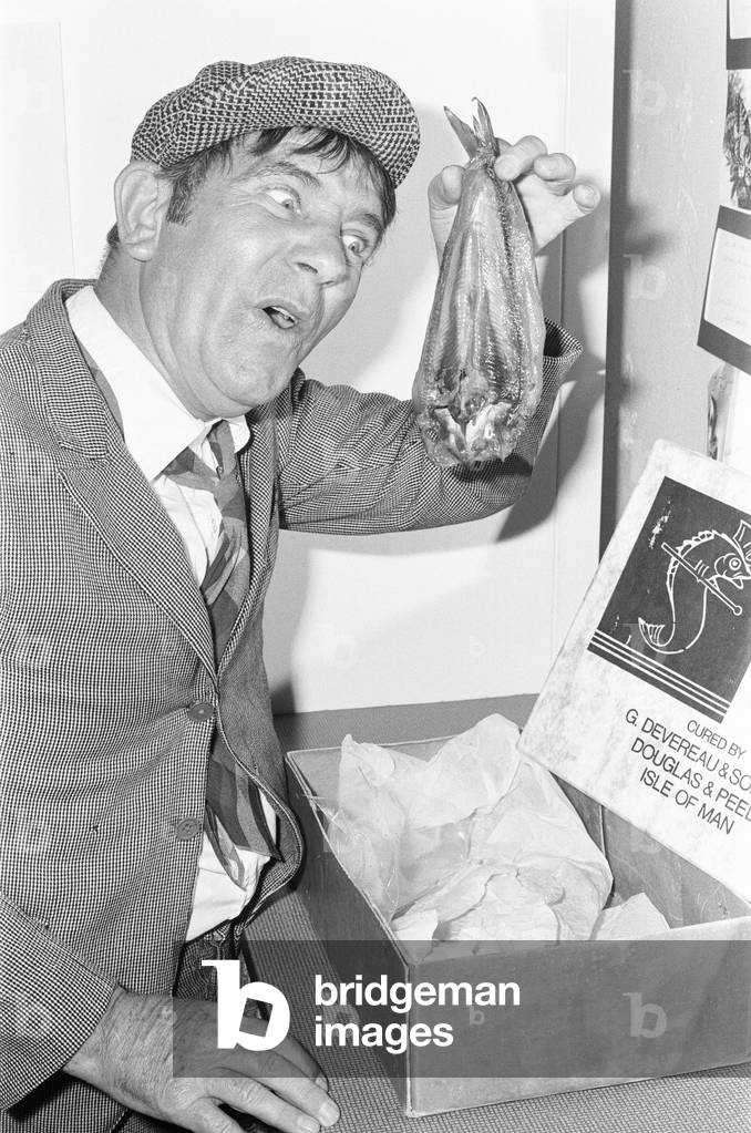 Comedian Norman Wisdom seen here in the persona of hapless on screen character Norman Pitkin in his dressing room in Douglas on the Isle of Man holding a kipper. 28th August 1977 (b/w photo)
