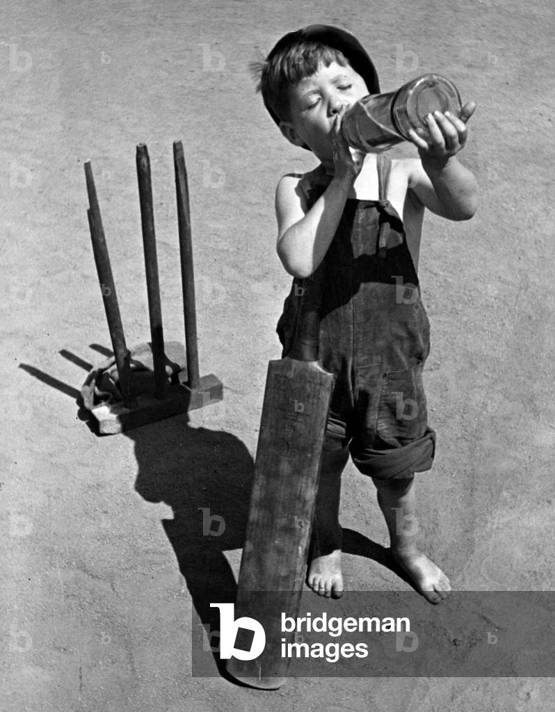 Five year old boy enjoys a refreshing drink from a bottle whilst playing cricket on a hot day in Shadwell.