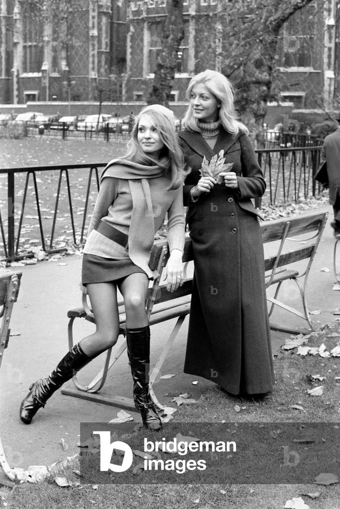 Model Blanche Webb wearing mini skirt and knee high boots with model Monica Hahn wearing a maxi coat as they pose together by a bench. 
November 1969
