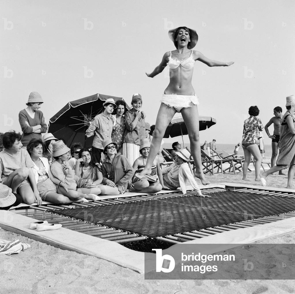 A model wearing bikini swimwear bounces on a trampoline on a beach in the French Riviera. 2nd August 1961 (b/w photo)