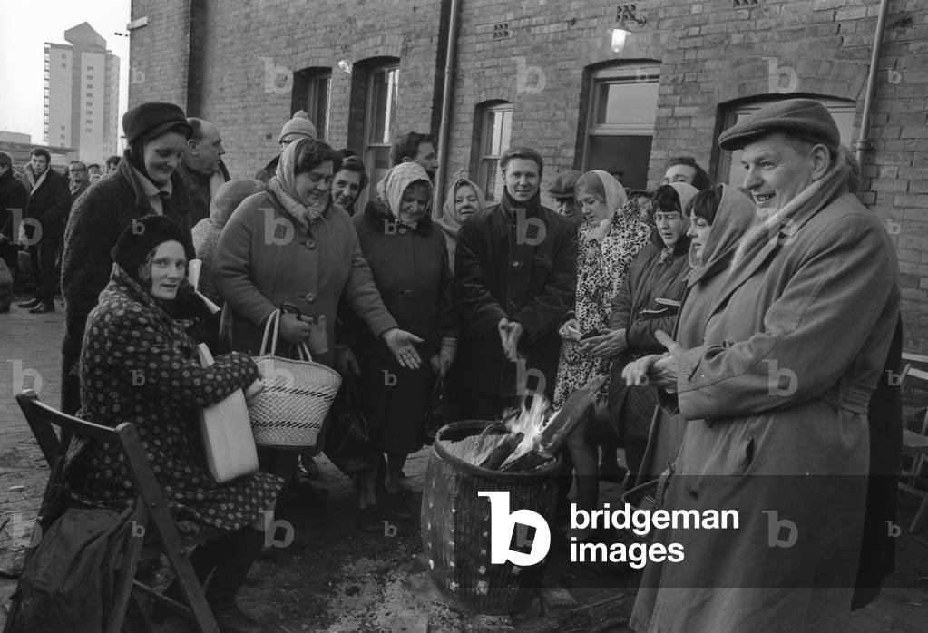 Only the brave will forgo the warmth of their beds to stay up all night in temperatures that fell to 26 degrees F. Nearly 60 did just this last night in their bid to book chalets at the Co-op. private holiday camp at Rhyl, North Wales. By sunrise a long queue was forming outside the booking office in King Street, Coventry. A wash tub brazier filled with logs, pictured here, helped to keep out the cold. 2nd February 1969 (b/w photo)