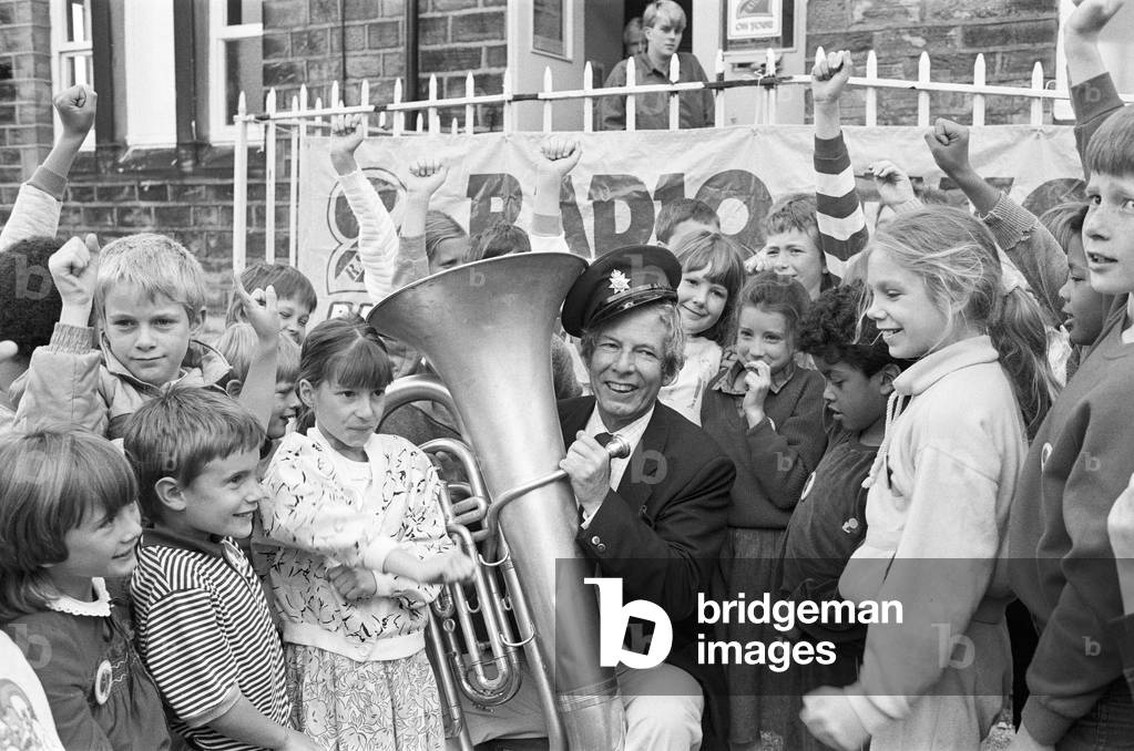 Cor Blimey - it's Derek Jameson... Radio 2 star Derek, complete with Cockney drawl and infectious chuckle delighted these children at Hinchliffe Mill Junior and Infants School, Holmbridge, with attempts at tuba playing this morning. In response to a letter from 10-year-old pupil Sarah Walker, Derek broadcast today's Radio 2 breakfast show from the school hall. 15th September 1987 (b/w photo)