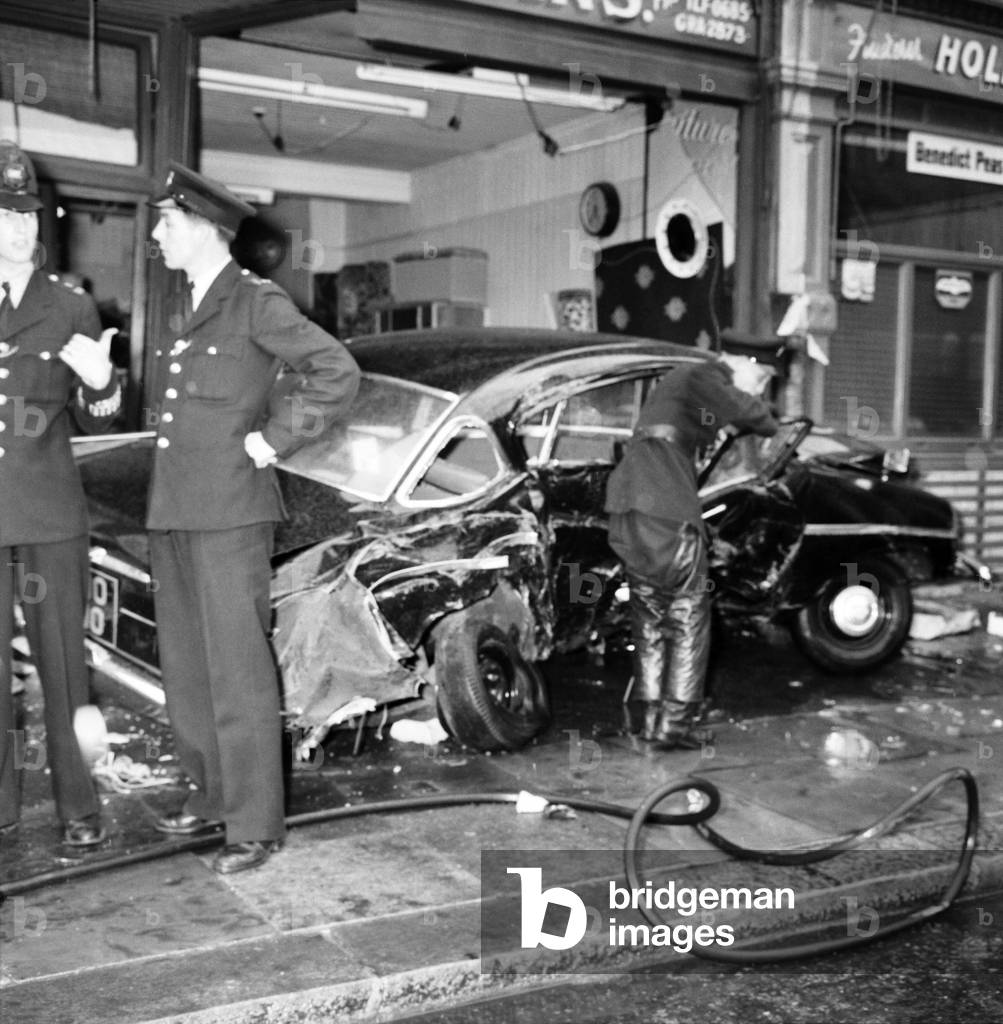 Police and firemen inspect the damage following a crash at Green Street, Upton Park. were 2 cars and a bus were involved in a collision. The bus ended up in the front of a shop . June 1960