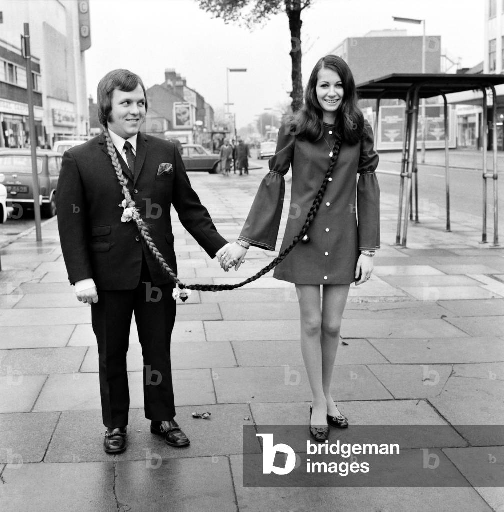 Ian Newman (19) a car salesman in Hounslow was getting worried as every time he got a new girlfriend and took her to dances other men who took a fancy to his girl would steal off with her and so Ian was left girl-less. But now he has the solution provided for him by hairdresser Len Pountney who has designed a long pigtail from Ian's hair that connects to Gloria Wootton (15) a florist, so that now everywhere the couple goes they are linked together. Ian Newman and Gloria Wootton linked in the high street today. November 1969
