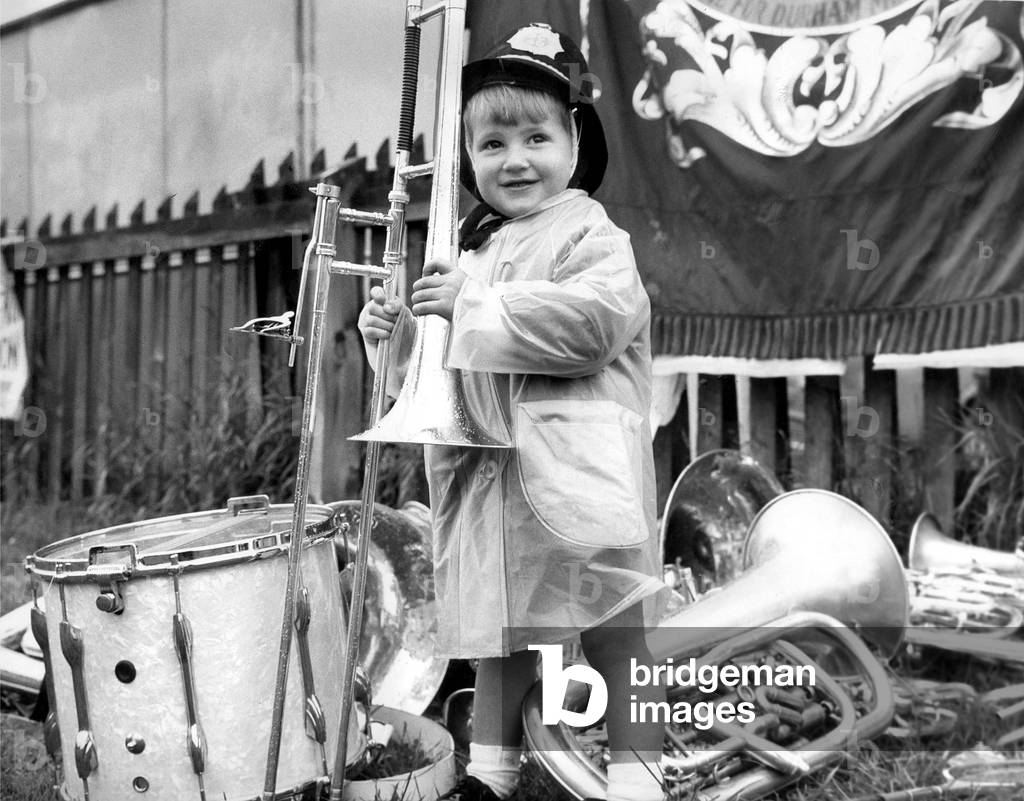 Durham Miners Gala - The rain may pour down, but 2 year old Crawford Cook of Murton Colliery stands his ground complete with policemen's helmet to watch over the band's instrument's, c. 1960
