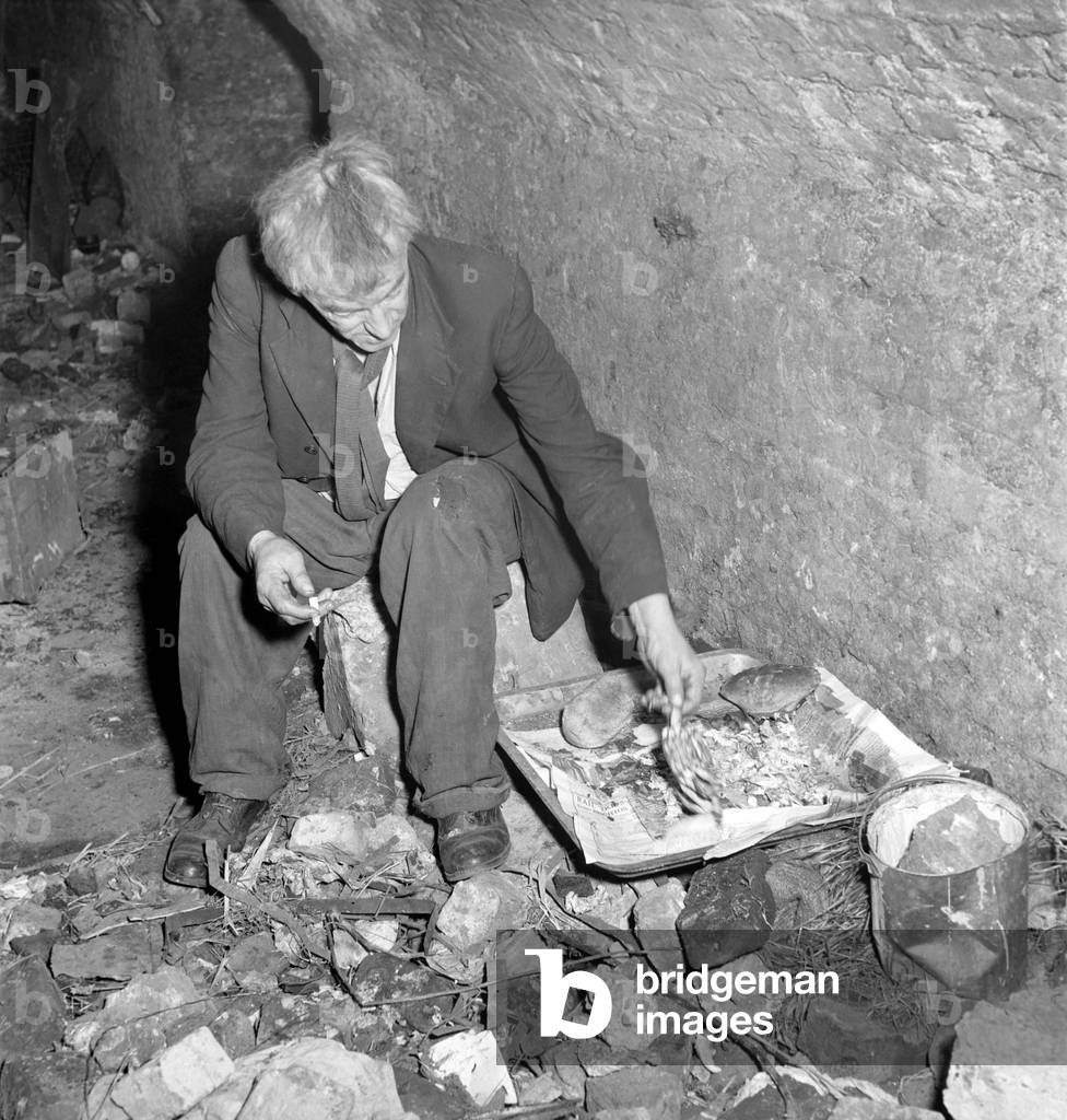 One of two homeless gentlemen who live in the cellar of a bombed out building close to Tower Bridge, seen here gather lunch together, 1953 (b/w photo)