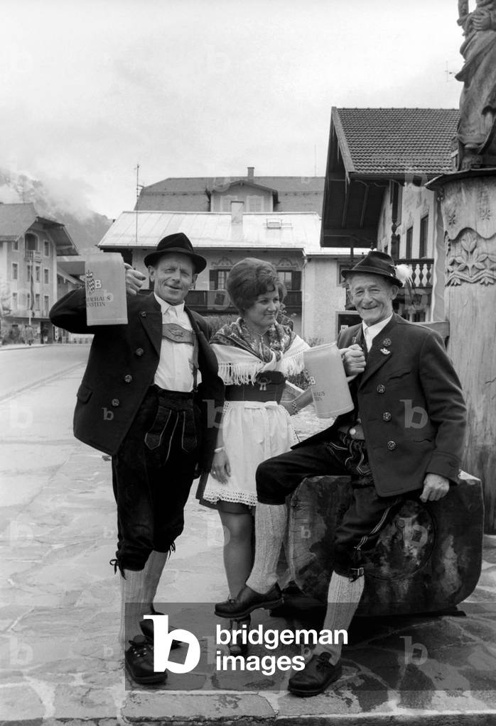 Chris Ward with people from the village of Ruhpolding, wearing their national costume and with three litre beer tankards in Munich, West Germany, May 1975 (b/w photo)