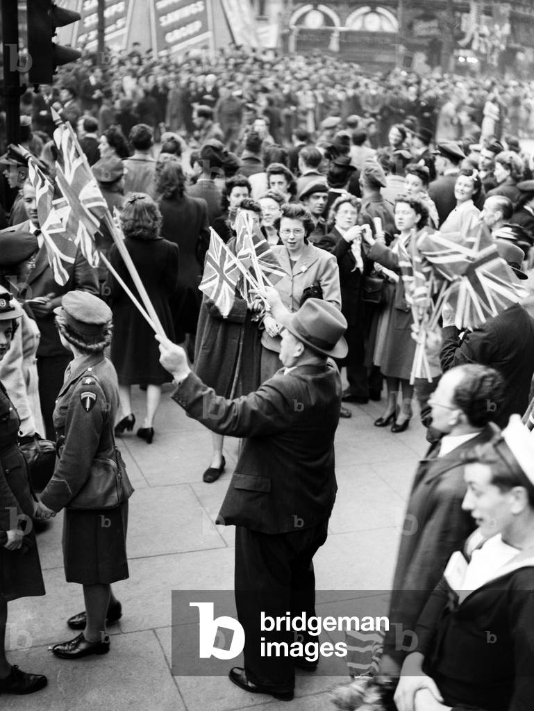 VE Day celebrations in London at the end of the Second World War. Huge crowds gathered around Piccadilly Circus during the celebrations. A patriotic reveller waving his collection of Union Jack flags surrounded by servicemen and women as well as civilians., 8th May 1945 (b/w photo)