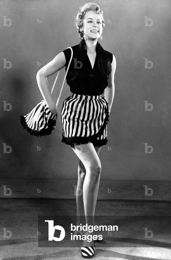 Woman dressed up for aday out at the beach, wearing sleveless top and striped skirt, carrying a matching handbag. 
April 1954