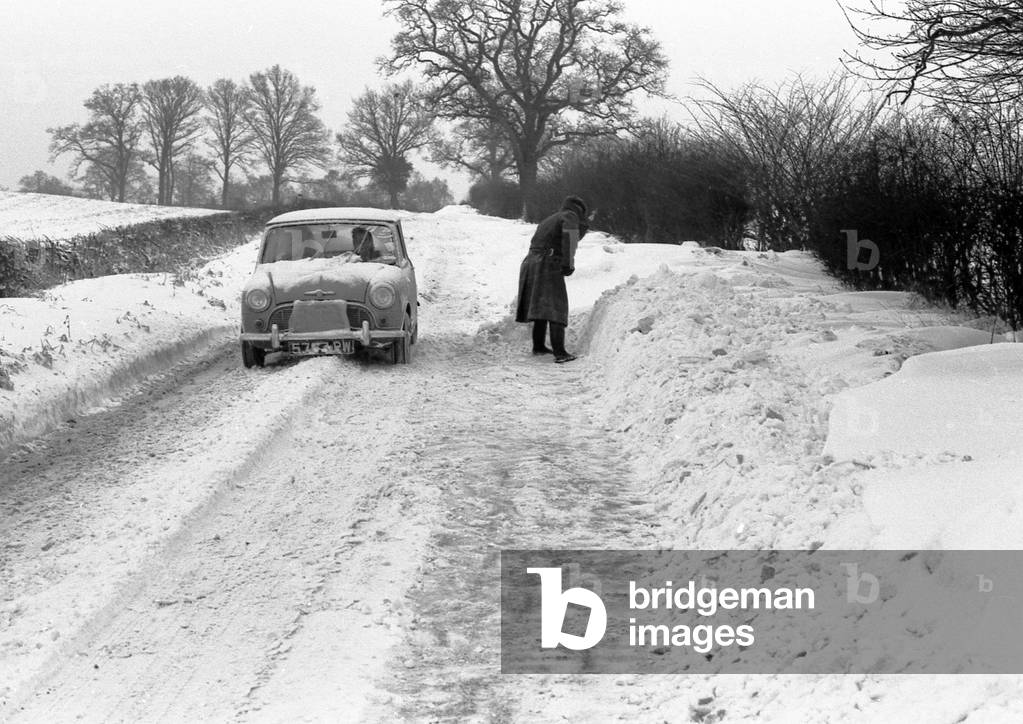 A mini car drives along a snowy lane in the countryside near Coventry whilst a man tries to clear away the snow. 3rd January 1963 (b/w photo)