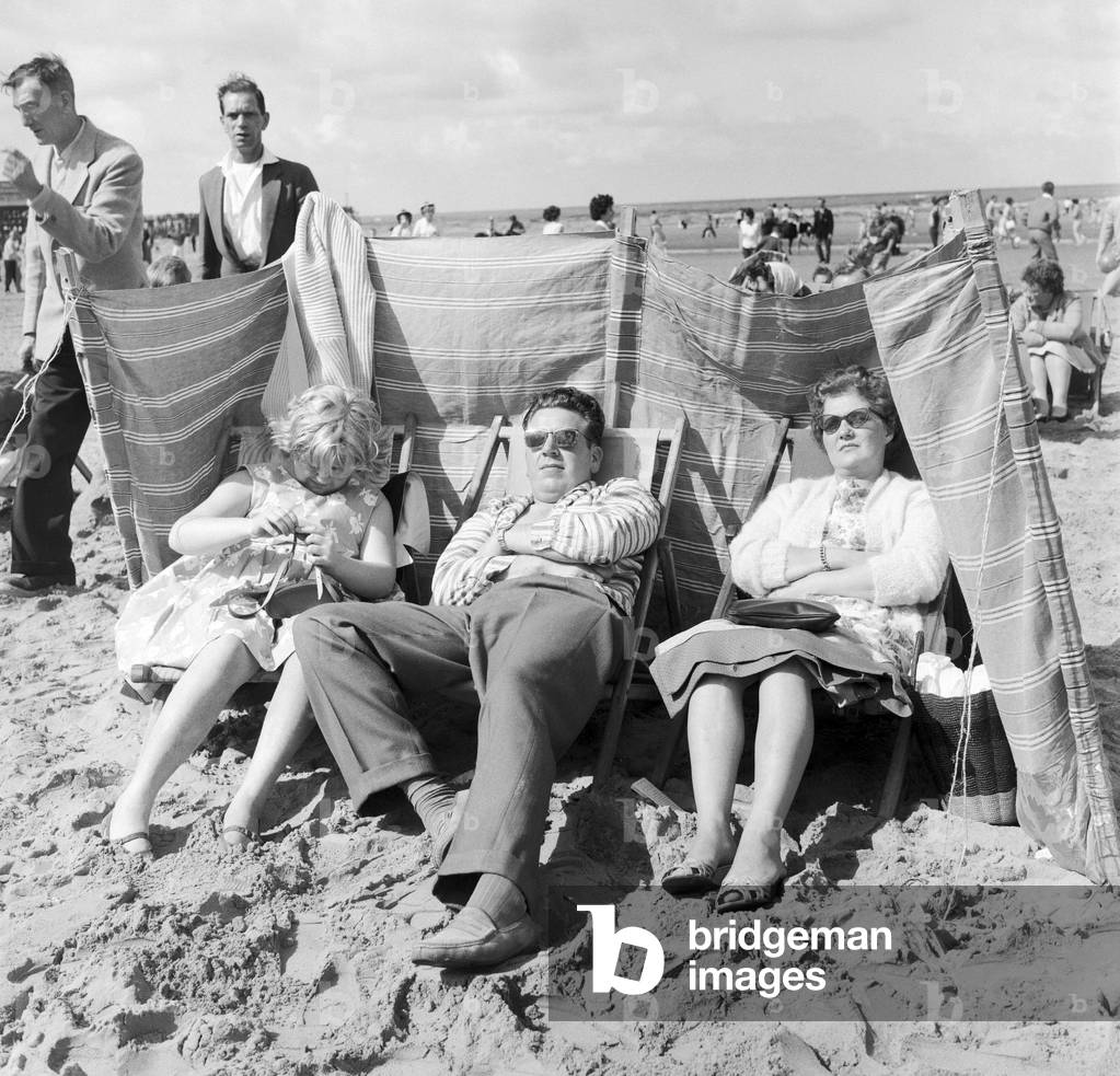 The Lemm family of Castleford, Yorkshire sit behind the protection of their windbreaker during a windy at Blackpool beach in the summer holidays. 6th August 1961 (b/w photo)