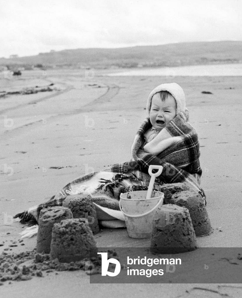 Poor Tracey isn't exactly having a ball-more of a bawl! Wrapped in blankets, her parents attempt to show her a good time building sand castles at Ayr Beach. 23rd June 1970 (b/w photo)