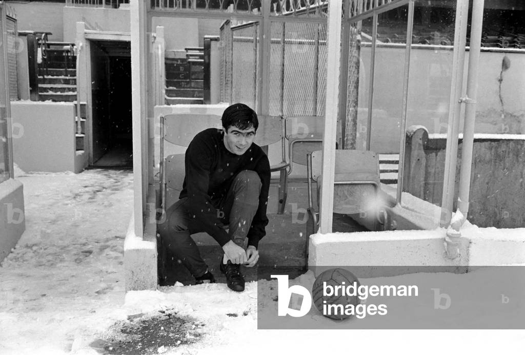 Arsenal's transfer lister forward, Bobby Gould, who was to have played for Arsenal reserves and West Ham reserves at Highbury, donned his track-suit and loosened up on the pitch for our cameraman, in very thick snow in front of a completely deserted stadium. Bobby Gould adjusts his boots before training. November 1969 (photo)