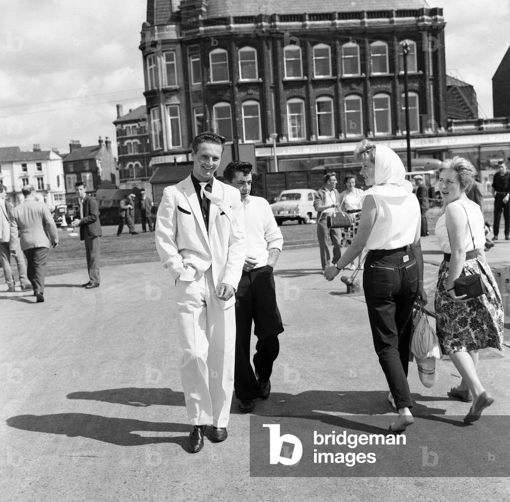 A new craze - Fancy suits worn by trawler fishermen at Lowestoft. Hylton Brighty, aged 16, wearing a white suit trimmed with black, watching him with interest are Mary Kampkin, 18, of Colindale (with headscarf) and Linda Suthers, of Kingsbury, who are both on holiday in Suffolk. 19th July 1961 (b/w photo)