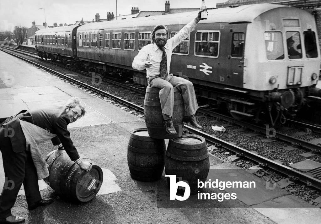Peter Gill (pushing barrel) and Dave Young (sitting on barrels) specialising in wine a barrel beer from their off licence in West Jesmond Station on 30th March 1976 (b/w photo)