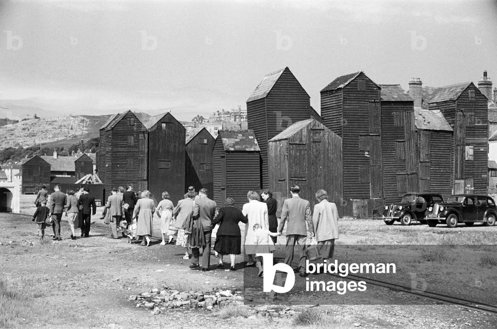 Holiday scenes. in and around Hastings, East Sussex.. Tourist walk along the seafront past the wooden sheds that holding the fishing nets of the local fishermen. June 1952 (b/w photo)