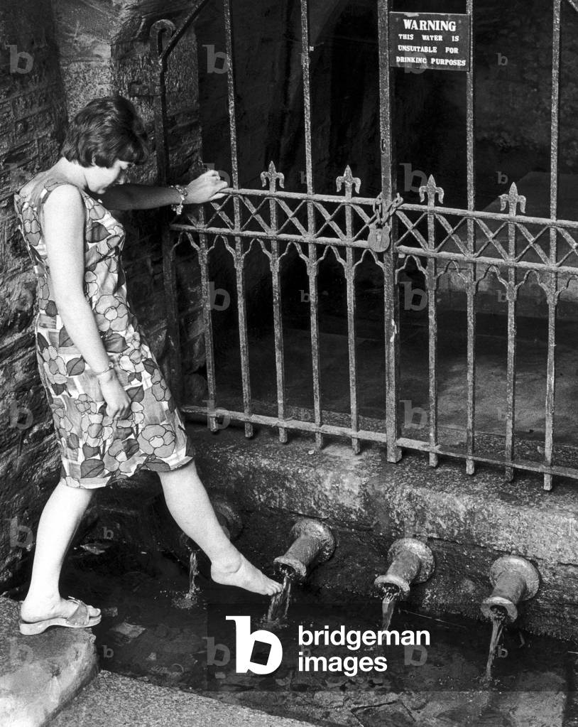 Heather Doney wets her foot at the pipe well in Well Lane at Liskeard, Cornwall. Heather was following an age-old custom in which water from any of the four pipes is supposed to have the mysterious property of making sure a girl will marry early. 29th September 1966 (b/w photo)