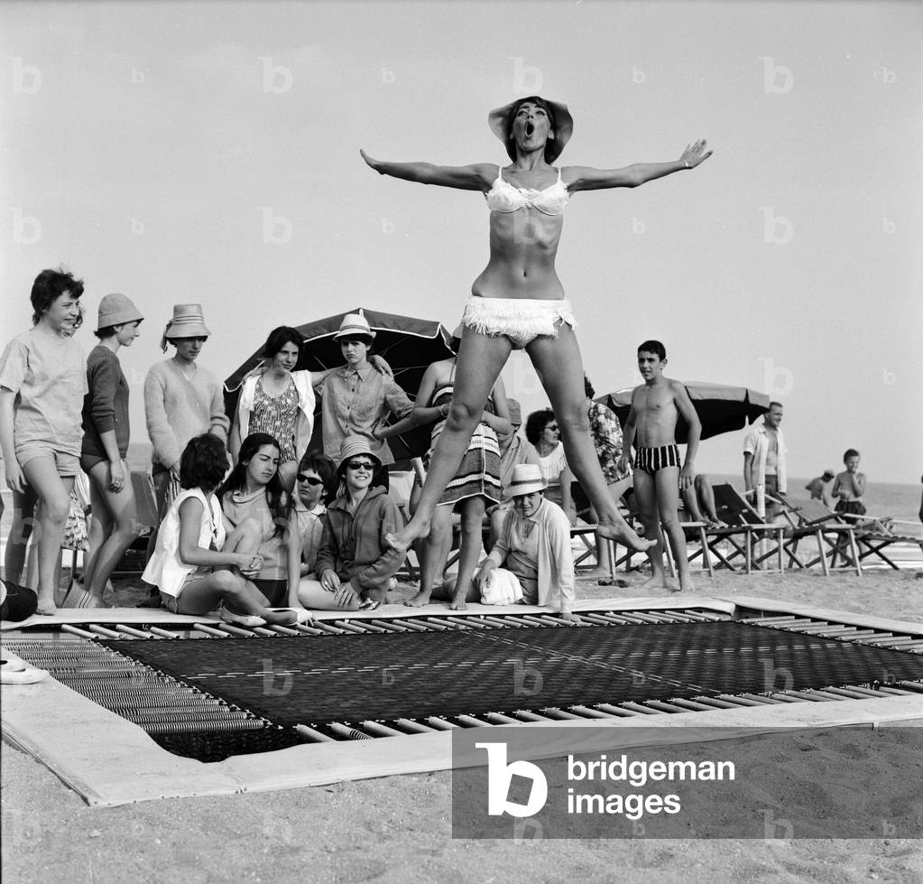 A model wearing bikini swimwear bounces on a trampoline on a beach in the French Riviera. 2nd August 1961 (b/w photo)
