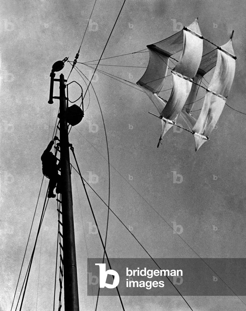 A seaman up the main mast adjusting the huge kite that carries a slender cable and make an effective barrage against enemy aircraft. Each ship in the convoy carries one of these. December 1940 P011670
