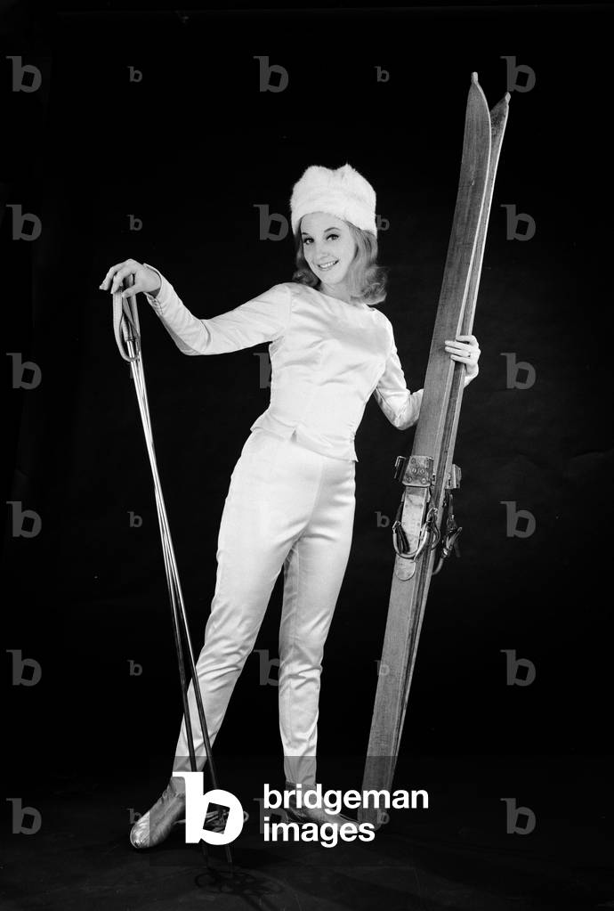 Woman in ski outfit with skis and poles, c. 1964 (b/w photo)