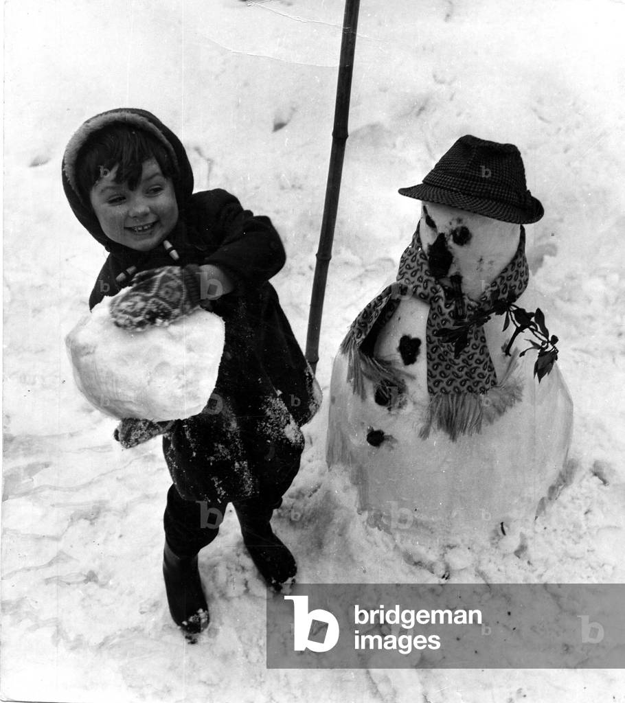 A snowman dressed in dad's hat and scarf gets a pounding from this youngster enjoying his time in the snow, Cardiff, 1963 (b/w photo)