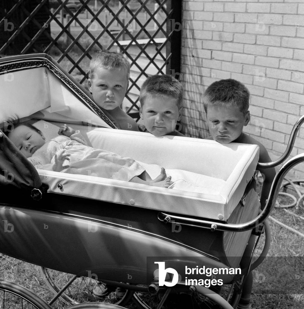 Three gloomy-faced triplet brothers aleaning over the side of a pram in the garden of their home at their new born baby sister. 
June 1960
