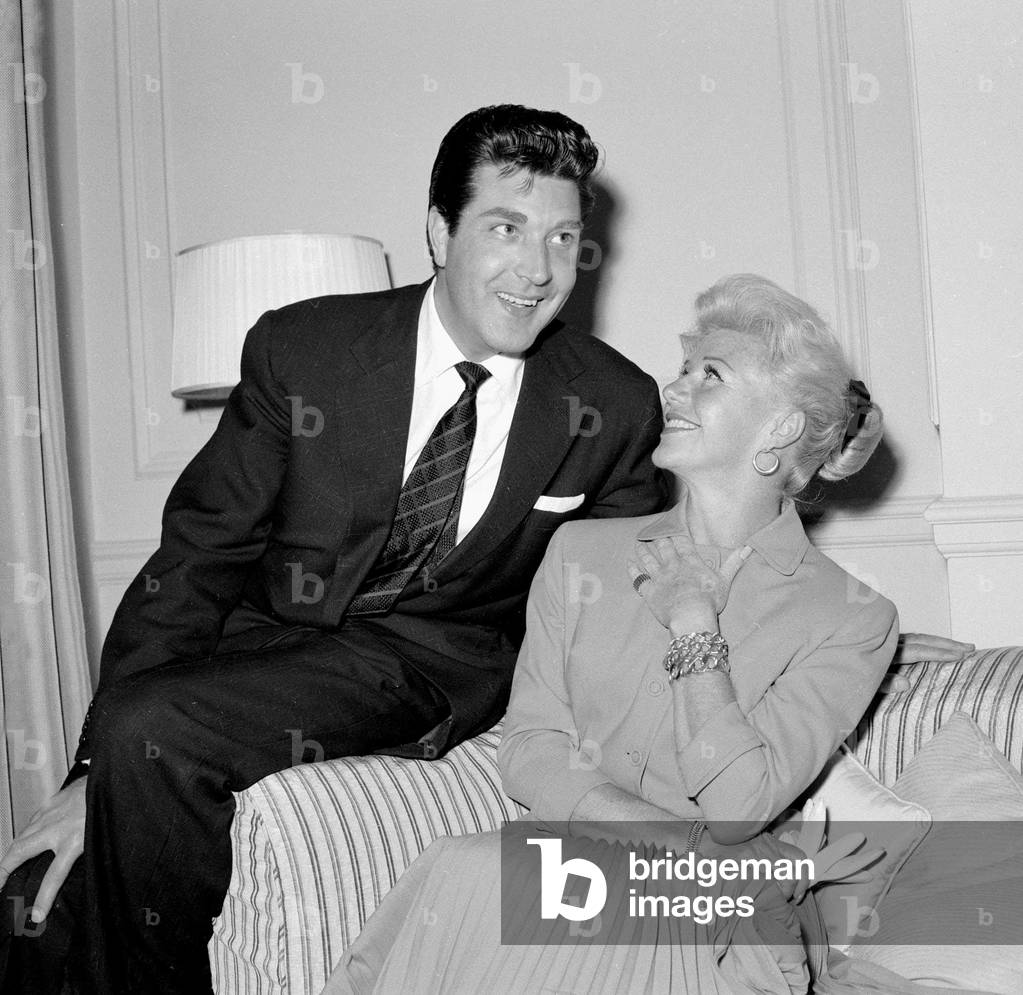 Ginger Rogers sitting on a couch laughing with David Hughes May 1959 (b/w photo)