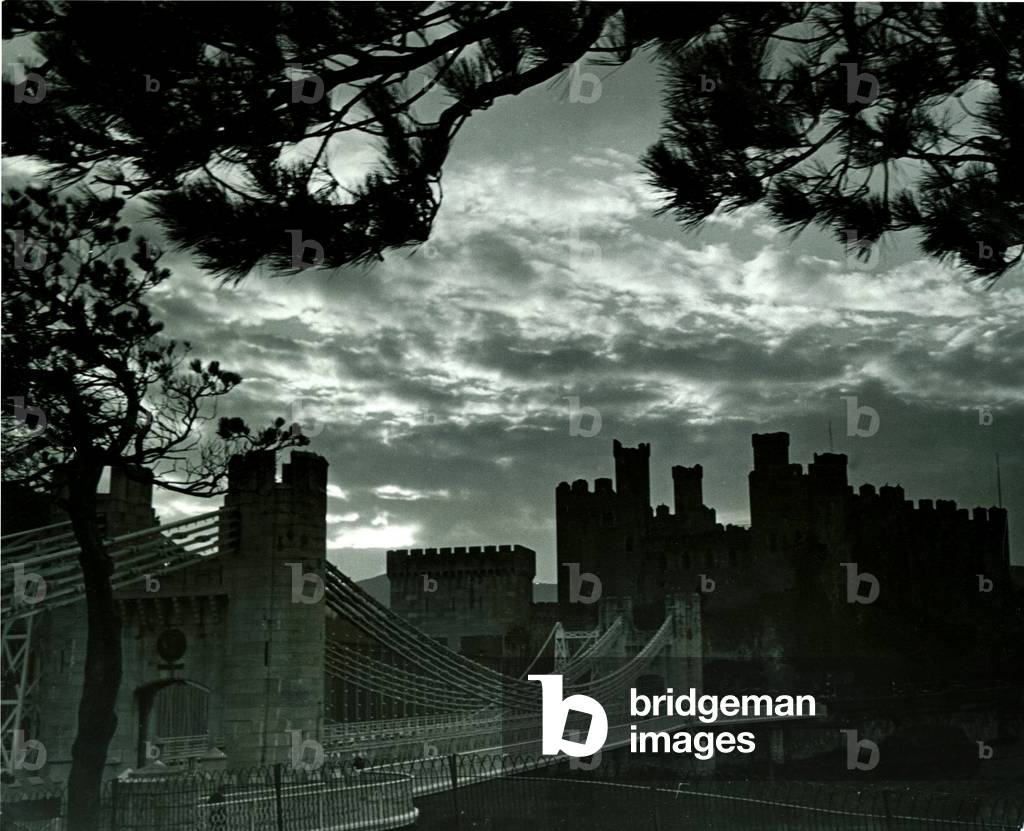 Night hides the scars of seven centuries, and in this picture Conway castle might still be magnificently defending the historic town of Conway one of Edward 1st chief bastions in the wars of North Wales Landmark Sunset Suspension Bridge Clouds Night Scene March 1942 (b/w photo)