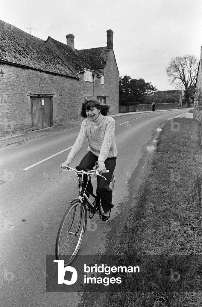 Pam Ayres riding her bicycle near her home in a village in Oxfordshire. 12th May 1977 (b/w photo)