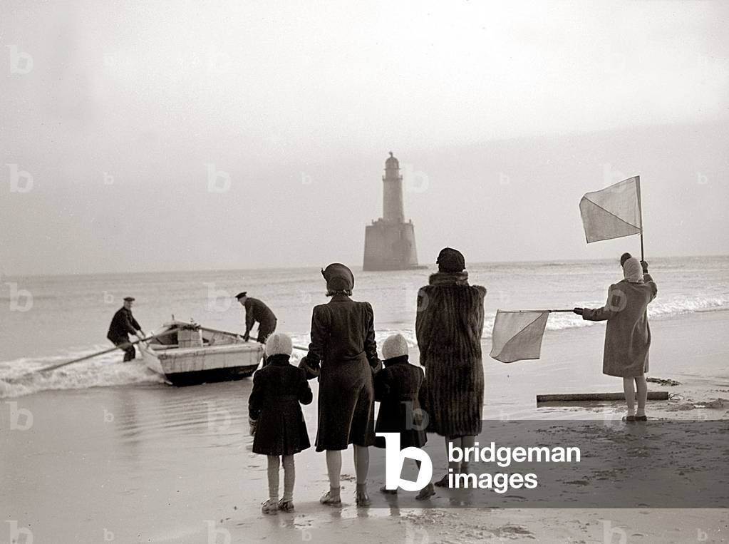 The families of the Rattray Head lighthouse keepers semaphore their loved ones as Old Alick prepares to take out Christmas gifts to the lighthouse, December 1943 (b/w photo)