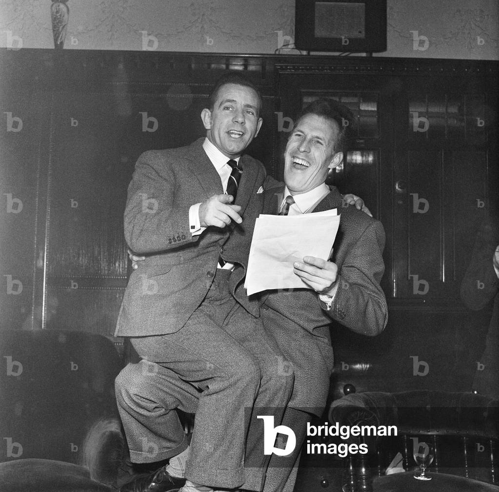 Norman Wisdom sits on Bruce Forsyth's knee and looks at the script for a pantomime which Norman is to appear in at christmas time. November 1959 (b/w photo)