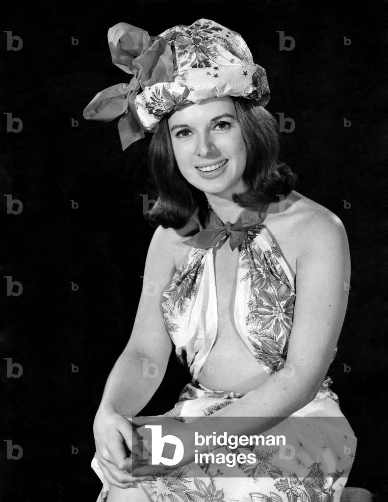 Reveille Fashions 1964: Jacqueline Waller modelling a halter top dress with a winter print scene and matching hat, December 1964 (b/w photo)